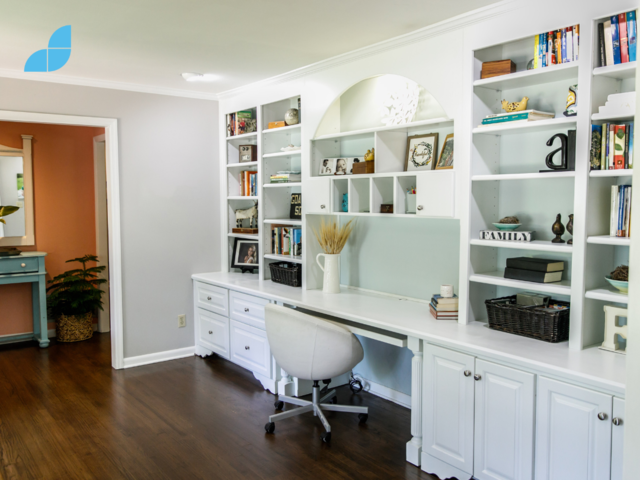 A office design with built in bookcases and cabinets, dark wood flooring and white furniture. Several plants and decoration can be seen along the shelves. 